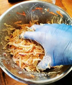 En hand med blå handske blandar coleslaw i en metallskål.