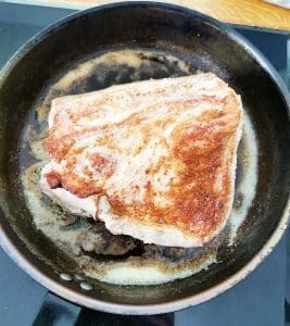 En bit fläskkarré som steks i en svart stekpanna med smör.