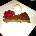 En skiva chokladkaka med nötter, serverad med grädde och hallon på en vit tallrik.