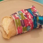 Semmelwrap med grädde inlindad i färgglad pappersförpackning på en tallrik.