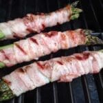 Tre bitar sparris inlindade i bacon som grillas på en grill.