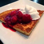 En bit tjockpannkaka med hallonsylt och vispad grädde på en vit tallrik.