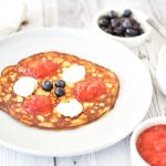 En pannkaka med blåbär och jordgubbssylt på en tallrik, med grädde i skålar bredvid.