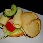 En sandwich med skivor av ost, grönsaker och en rund brödskiva på en tallrik.