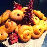 En hög med lussekatter placerade på en träskärbräda med små röda paket och en julgran.