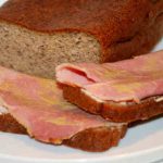 En skiva vörtbröd med skivor av kött på en tallrik, med en hel limpa vörtbröd i bakgrunden.