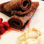 Två chokladcrêpes rullade och strödda med socker, serverade med hallon och grädde.