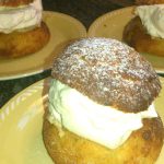 Tre semlor med vispad grädde på tallrikar, med pudrat socker på toppen.