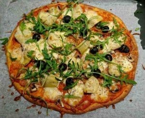 En pizza med ost, svamp, oliver, kronärtskocka och ruccola på en bakplåt.