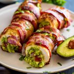 Guacamolefylld kycklingfilé inlindad i bacon på en tallrik med avokado.