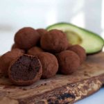 Avokadochokladbollar rullade i kakao på en träskärbräda med en halverad avokado i bakgrunden.