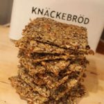 Staplat knäckebröd med frön och nötter på en skärbräda, med en burk i bakgrunden.