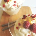Två skålar med citronmousse toppade med hallon och dekoration på en träskärbräda.