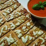 Krispiga brödsticks med smält ost på en skärbräda bredvid en skål med tomatsås.