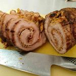 Skivad köttfärsrulle med kryddor på en skärbräda med en kniv bredvid.