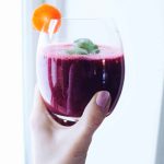 En hand som håller ett glas med röd smoothie, dekorerad med en skiva morot och basilika.