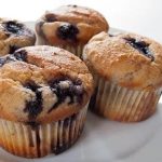 Fyra blåbärsmuffins med gyllenbrun yta placerade på en vit tallrik.