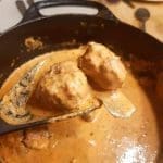 Köttbullar i en gräddig sås som lyfts med en slev från en svart gryta.