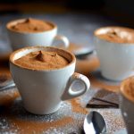 Fyra vita kaffekoppar med chokladpulver på toppen, placerade på en yta med chokladbitar.