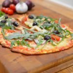 En pizza med tomater, oliver och ruccola på en träskärbräda.