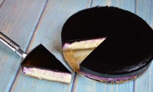 En blåbärs cheesecake med en spegelglasyr, en bit skuren och en serveringsspade bredvid.