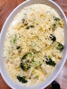 En gratäng med broccoli, grönsaker och riven ost i en vit ugnsform.