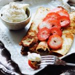 Två pannkakor fyllda med skivade jordgubbar och en skål med vispad grädde på en tallrik.