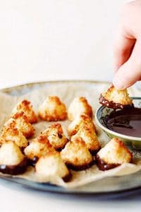 Kokostoppar på en tallrik med en hand som doppar en topp i en skål med smält choklad.