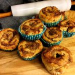En hög av kanelbullar i blå pappersformar på en träskiva med en kavel i bakgrunden.