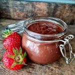 Glasburk med chokladkräm bredvid två färska jordgubbar på en träyta.