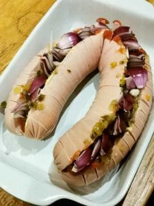 Två falukorvar med skivor av lök och grönsaker i en vit ugnsform.