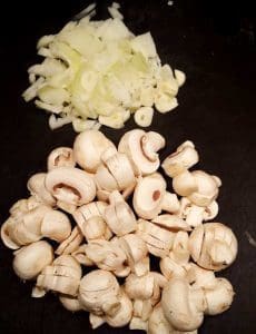 Skivade champinjoner och hackad lök på en svart skärbräda.
