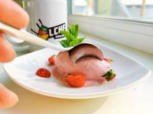 En skål med fryst yoghurt dekorerad med jordgubbar och mynta, med en sked i handen.