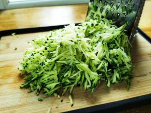 Rivna zucchinis i en hög på en skärbräda med ett rivjärn i bakgrunden.