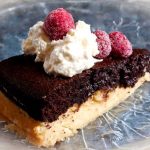 En bit chokladmazarinkaka med grädde och hallon på en glasfat.