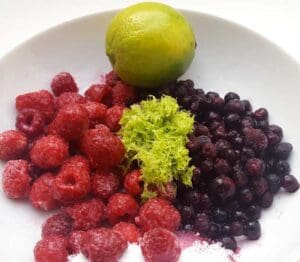 En tallrik med frysta hallon, blåbär, lime och limezest i en neutral miljö.