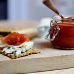 Glasburk med tomatmarmelad och knäckebröd med ost på en träskiva.