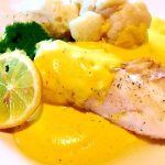 En tallrik med fisk täckt av citronsås, serverad med blomkål och en skiva citron.
