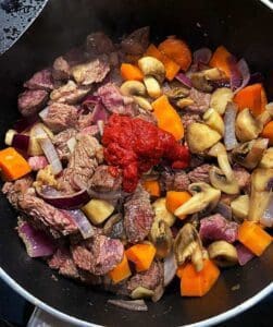 En gryta med kött, morötter, lök, svamp och tomatpuré i en svart kastrull.