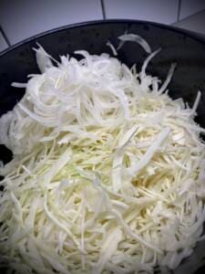 Riven vitkål och lök i en svart skål, med olika tjocklekar på strimlorna.