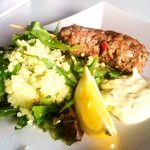 Lammfärsspett serverat med sallad, citronklyftor och en vit sås på en tallrik.