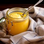 En glasburk med ghee och en sked på en grå och vit rutig duk.