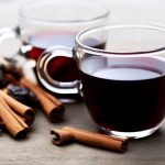 Två genomskinliga glas med starkvinsglögg och kanelstänger samt kryddor på en yta.