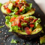 Halverad avokado fylld med bacon, tomat och sallad på en svart platta.