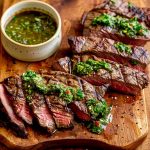 Skivor av grillat kött med chimichurri sås på en träskärbräda.