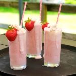 Tre glas med jordgubbsmilkshake toppade med grädde och jordgubbar, placerade på en bricka.