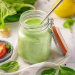 Grön basilika dressing i en glasburk med lock och sked, omgiven av färska ingredienser.
