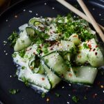 Krämig gurksallad med skivor av gurka, örter och sesamfrön på en svart tallrik.