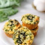 Tre äggmuffins med grönkål på en vit tallrik, med grönkål i bakgrunden.