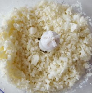 Riven blomkål i en matberedare med en vit kniv i mitten.